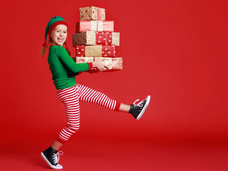 Child dressed as a holiday elf carrying a tall stack of wrapped gifts against a bright red background, featured for Vacaville Commons’ holiday season.