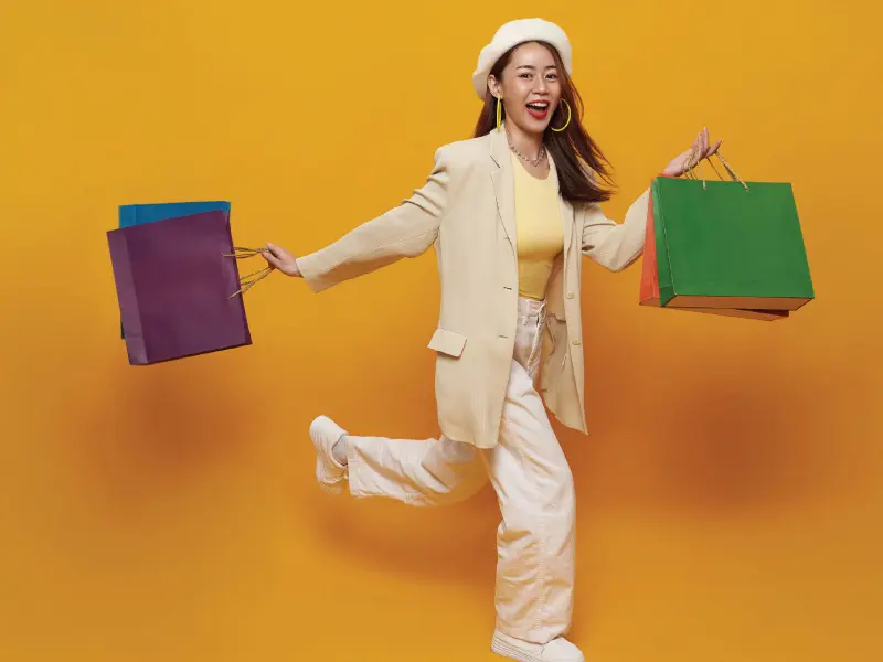 Woman with long brown hair, poofy white hat, tan jacket, pants, and soft yellow shirt holding shopping bags.